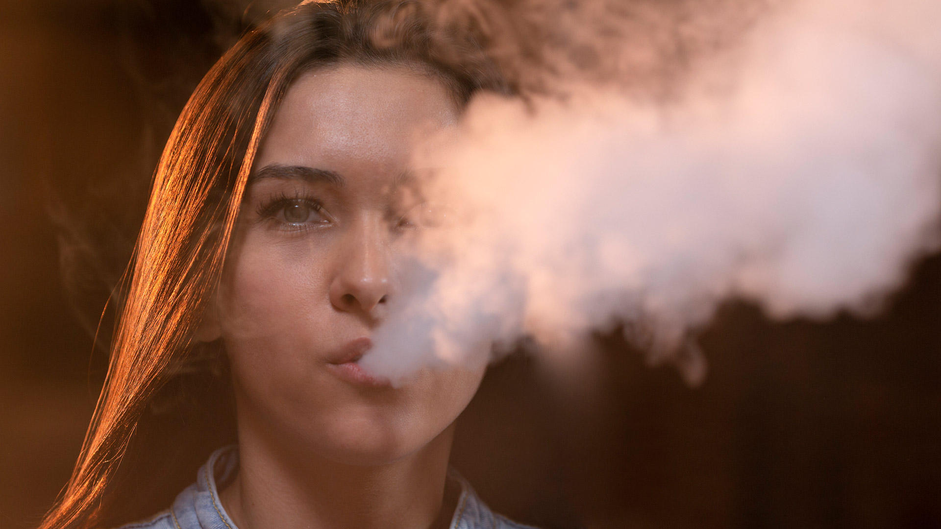 Young woman exhaling vapor, with a pensive expression, against a blurred indoor background.