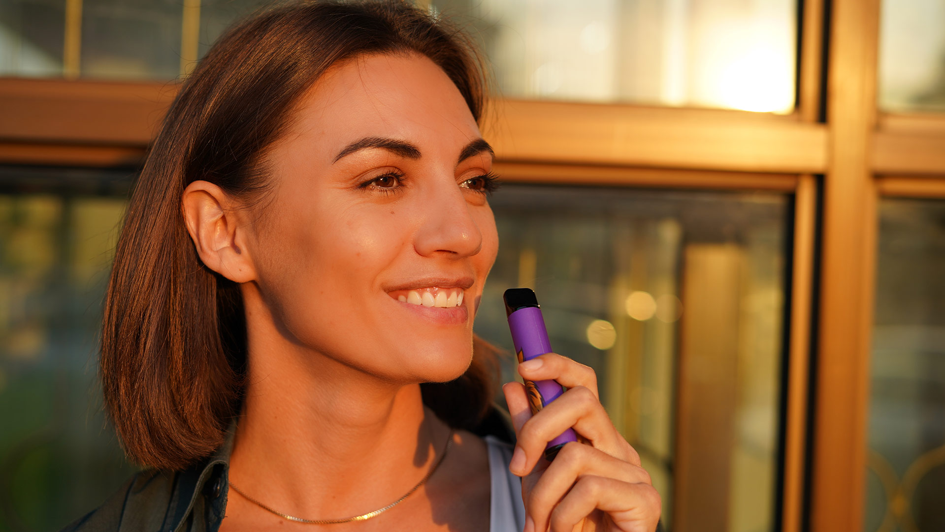 Smiling woman using a vape pen outdoors at sunset, depicting the lifestyle of reducing nicotine consumption through vaping.
