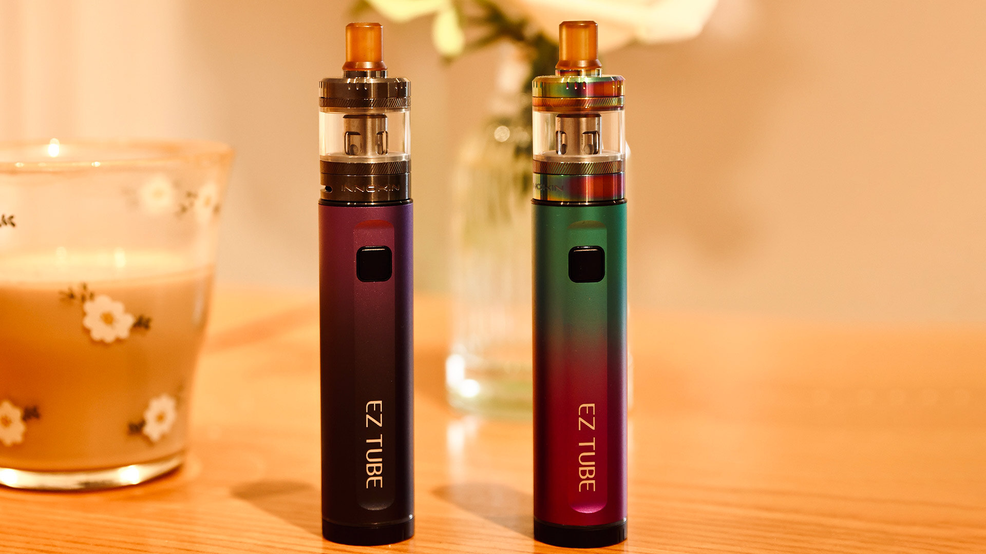 A pair of colorful EZ TUBE vape pens standing upright on a wooden table beside a floral-patterned glass, emphasizing a common issue—why a vape pen might not work after being charged—such as the device’s battery nearing the end of its lifespan.