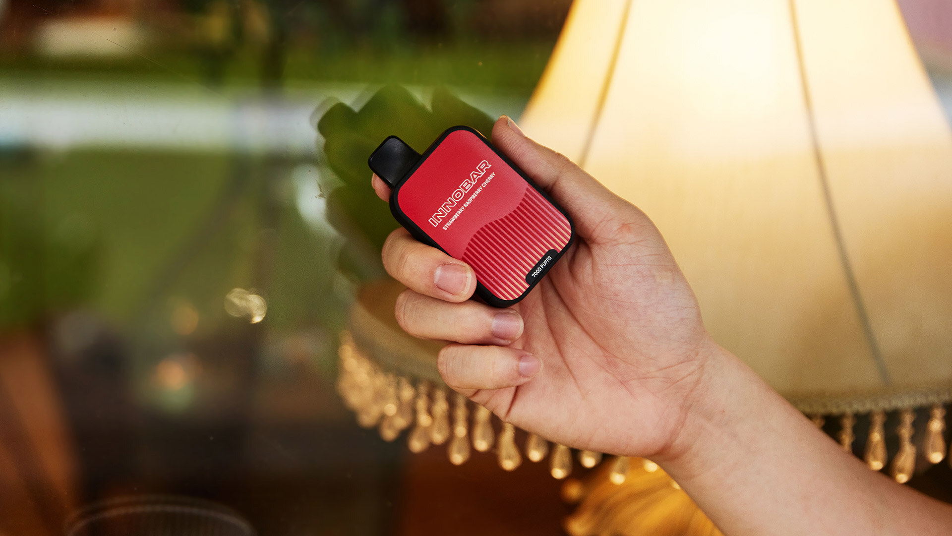 A hand holding a rechargeable disposable vape labeled INNOBAR and 7000 Puffs in front of a glass wall, with a lamp visible in the background.