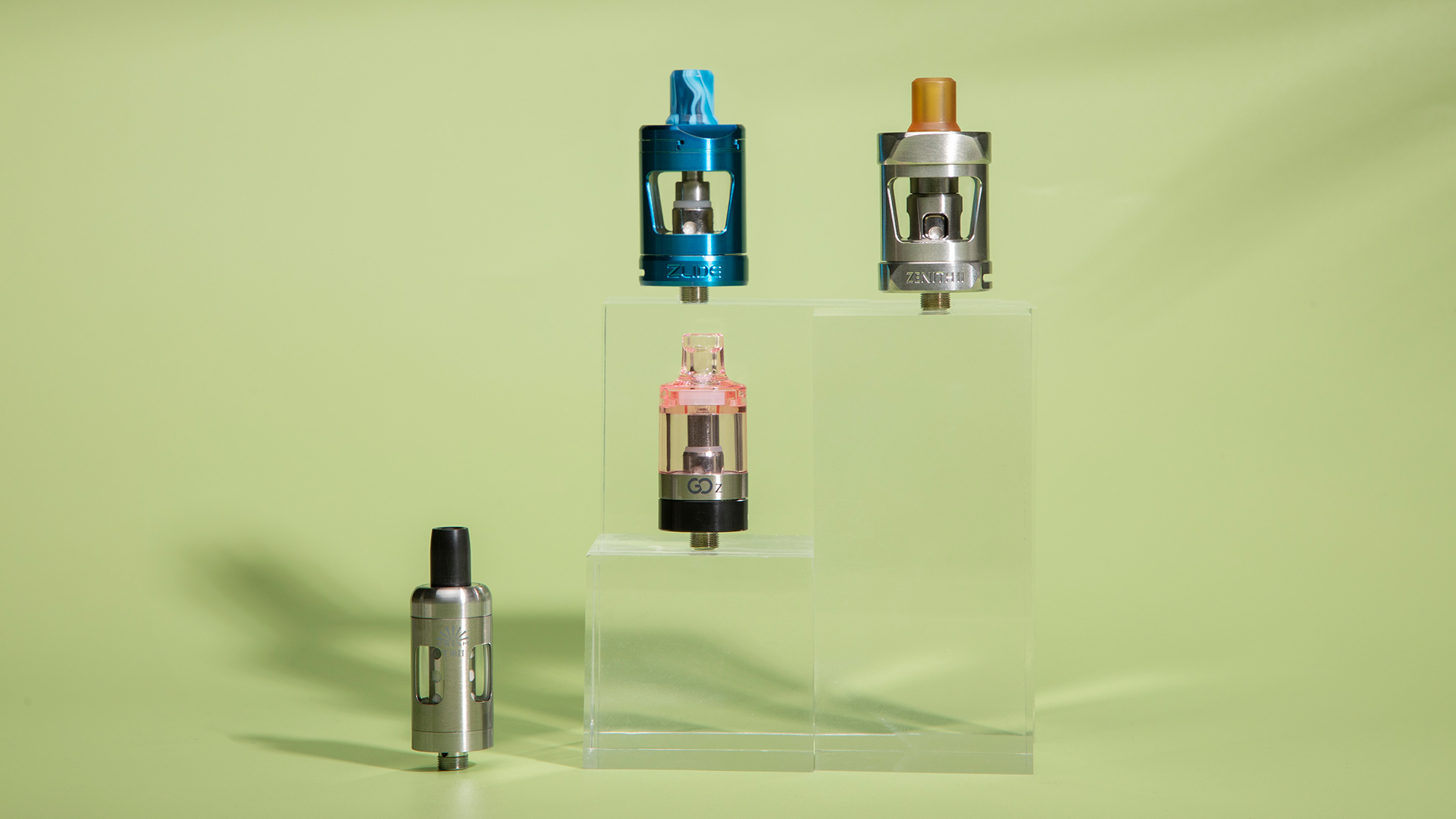 Four e-cigarette atomizers in varying colors and sizes, showcased on transparent stands against a light green background, potentially featuring MTL Tank designs.