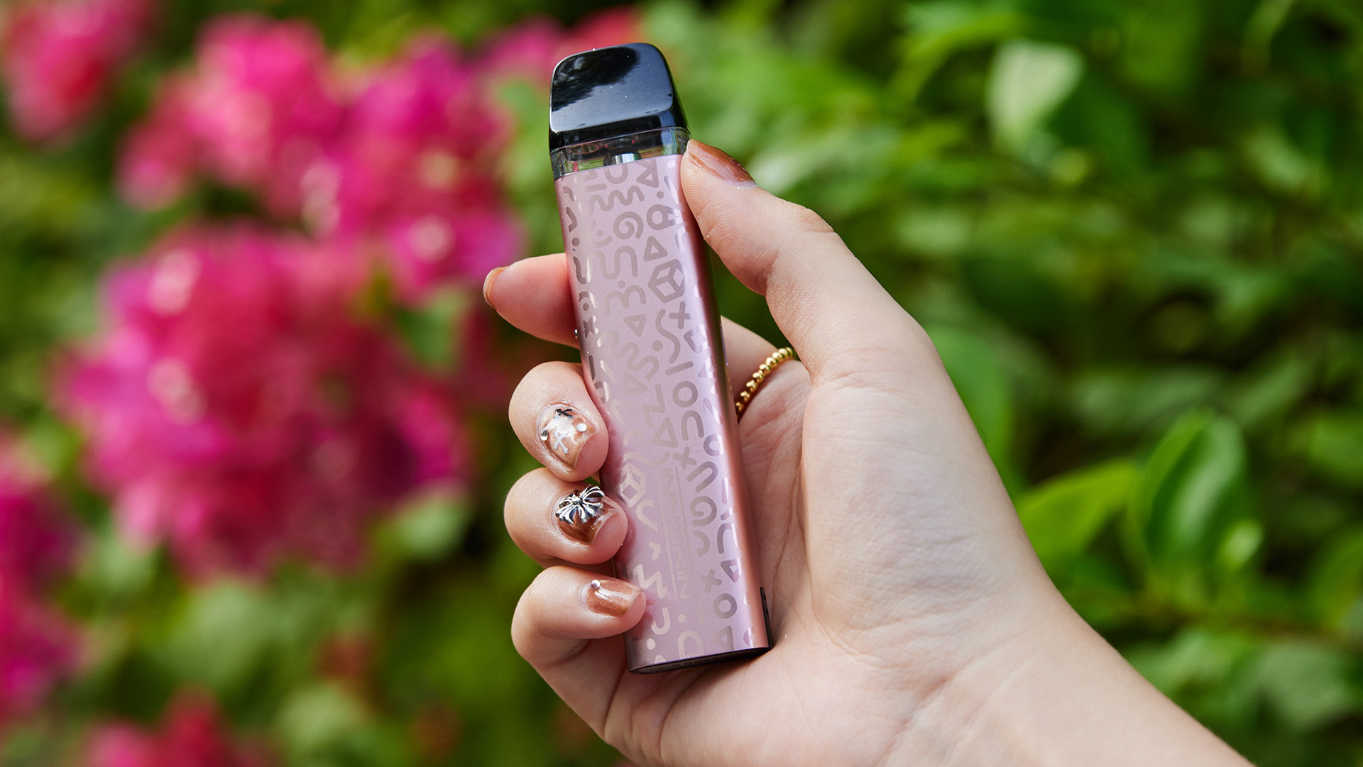 A hand holding the Innokin Trine SE pod vape in rose gold, adorned with a subtle pattern, against a backdrop of vibrant pink flowers.