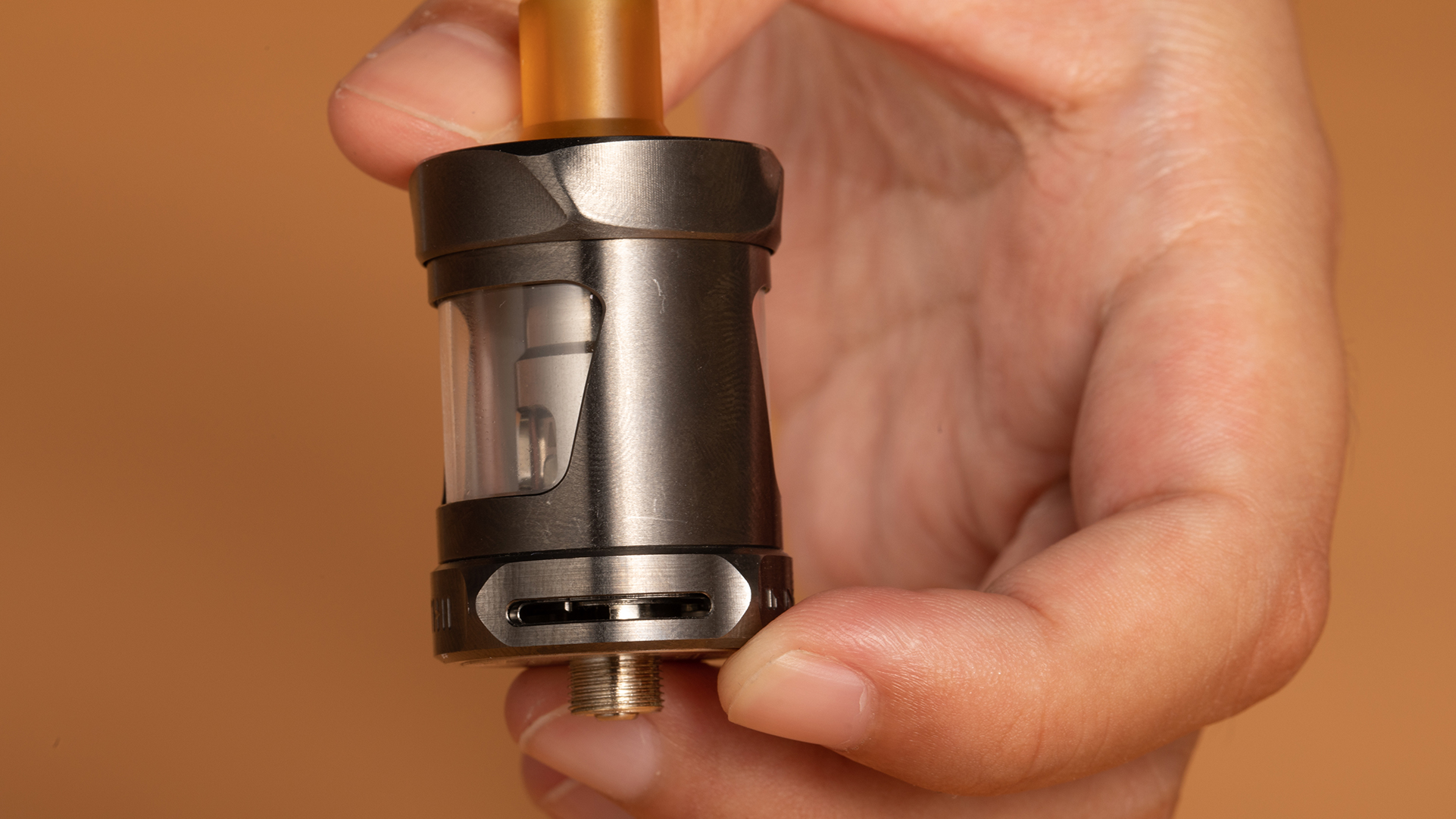 A close-up of a hand holding a vape tank with a visible coil inside, showing the bottom threading and connector. The image focuses on the tank as part of troubleshooting steps for fixing the 'No Atomizer' error if the tank or pod has a problem.