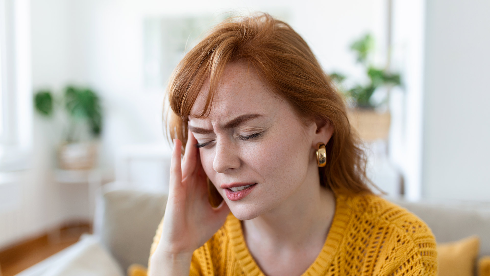 A redheaded woman in a yellow sweater grimaces and holds her head in discomfort, illustrating the physical strain and withdrawal symptoms associated with quitting nicotine before a drug test.