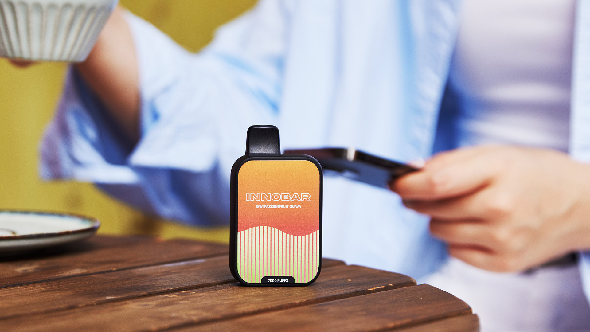A disposable vape device labeled INNOBAR and 7000 Puffs placed on a wooden surface, with a girl lifting a cup of coffee, suggesting a lasting and satisfying vaping experience.